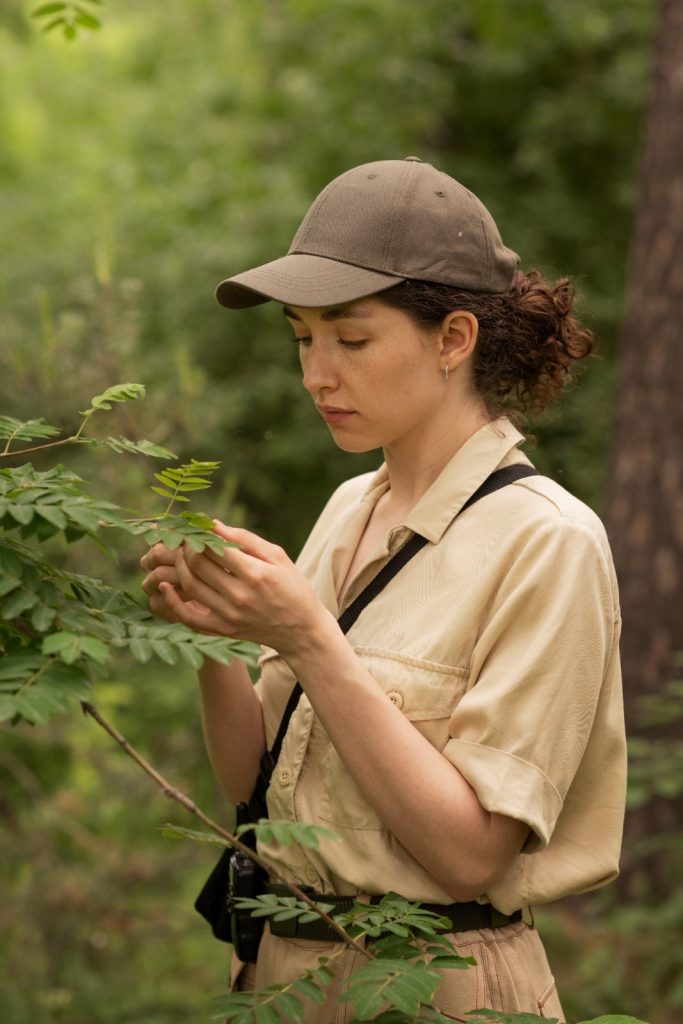 A woman in a beige shirt and cap examines a green plant in a lush forest, with a camera strap visible across her shoulder.